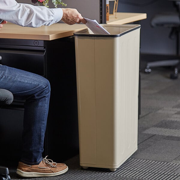 A man sitting at a desk in a corporate office and looking at a paper in a Rubbermaid beige steel trash can.