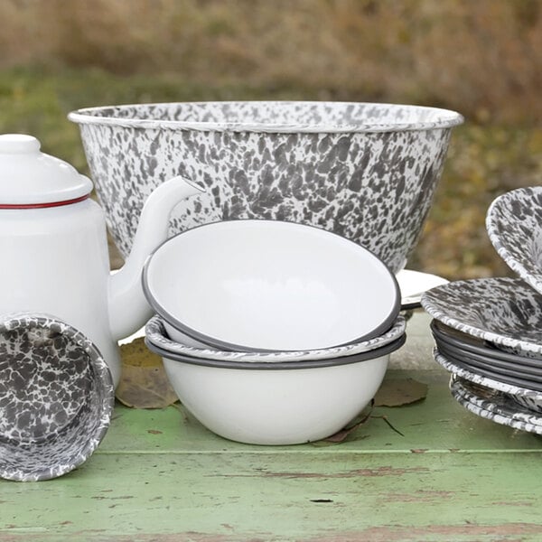A white enamelware bowl with a grey rim on a table with enamelware dishes and a teapot.