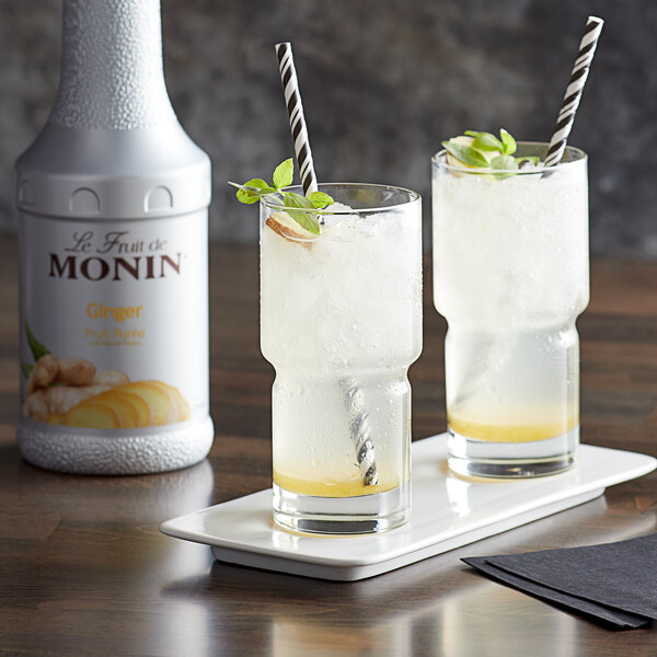 Two glasses of ginger lemonade with straws and mint leaves next to a bottle of Monin Ginger Fruit Puree.