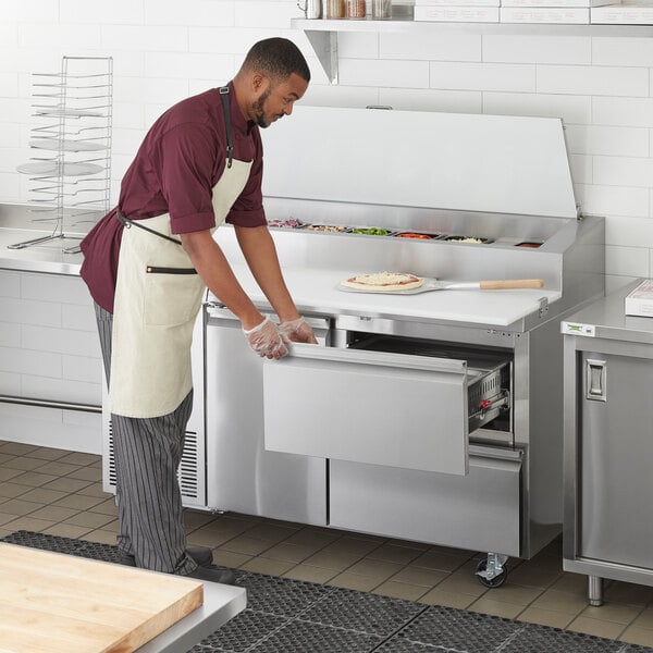 A man in an apron opening a drawer on a commercial pizza prep table.