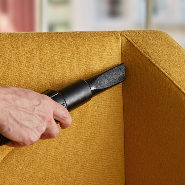 A hand holding a black Lavex crevice tool attached to a vacuum cleaner, vacuuming a yellow couch.