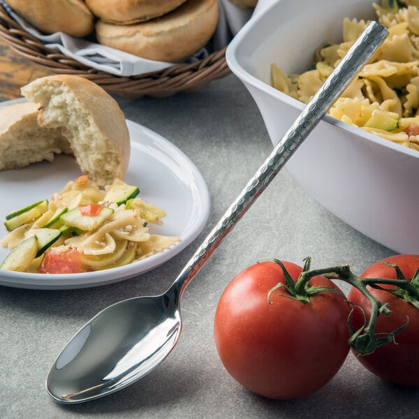 A Carlisle stainless steel serving spoon next to a plate of pasta and tomatoes.