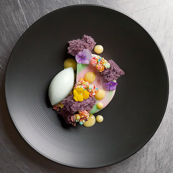 A dessert on a Front of the House semi-matte black porcelain plate with a flower on top.