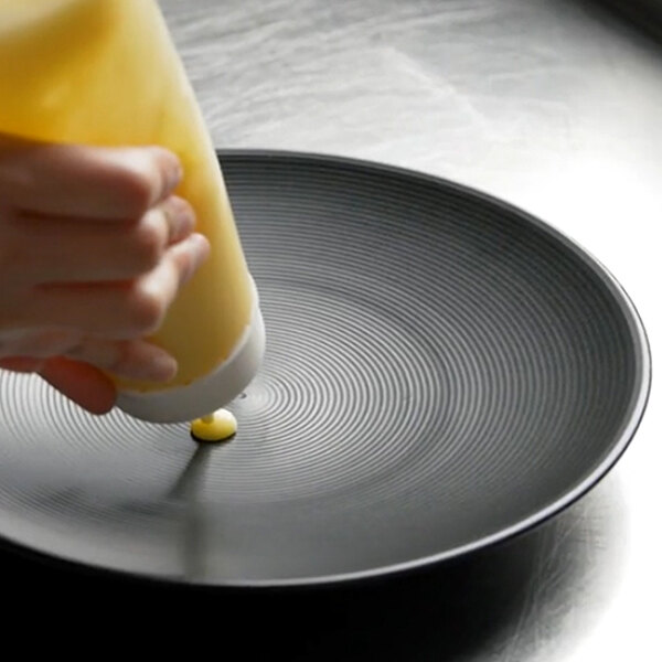A hand pouring yellow mustard onto a Front of the House Spiral Ink black porcelain plate.