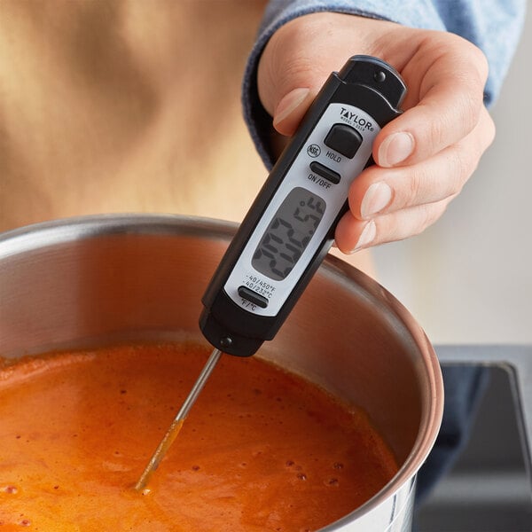 A hand holding a Taylor digital pocket probe thermometer to a pot of soup.