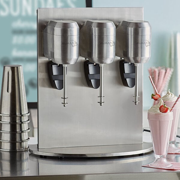 An AvaMix freestanding triple spindle drink mixer on a table with three silver containers filled with strawberry milkshakes.