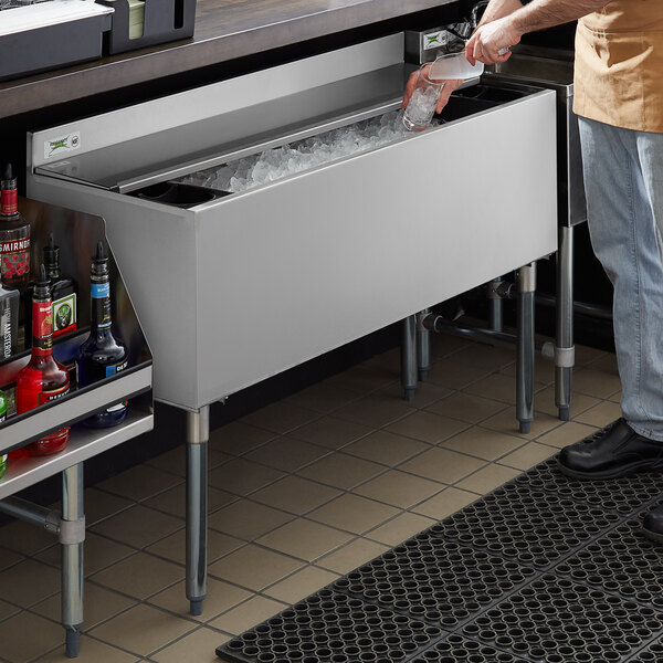 A man standing at a counter with a drink and a Regency underbar ice bin with sliding lid and bottle holders.