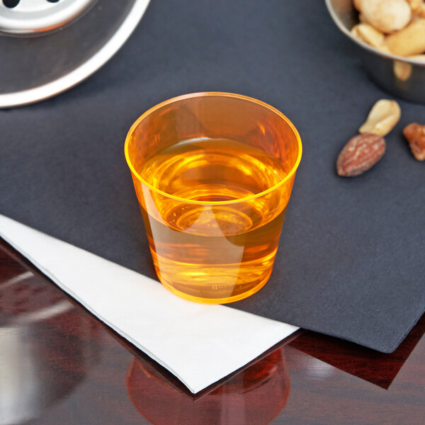 A Fineline Quenchers neon orange hard plastic shot cup filled with orange liquid on a table with nuts.