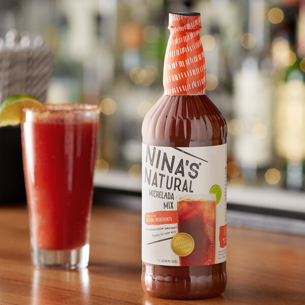 A bottle of Nina's Natural Michelada Mix with a white label on a table next to a glass of red liquid.