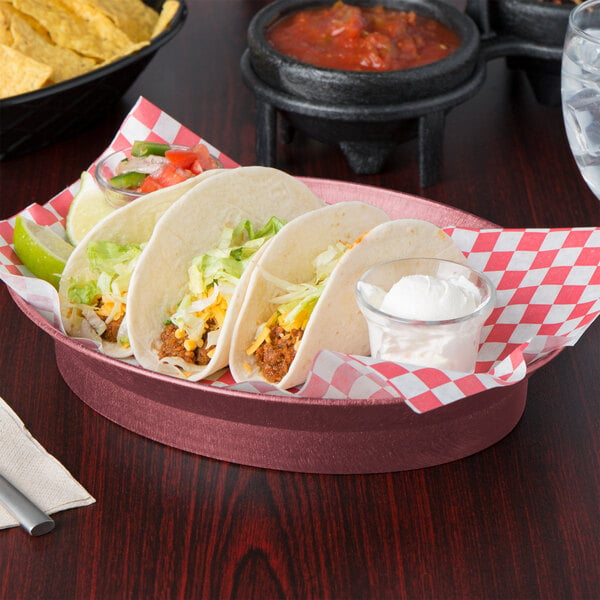 An oval raspberry deli server filled with tacos and a bowl of chips on a table with a red and white checkered napkin.