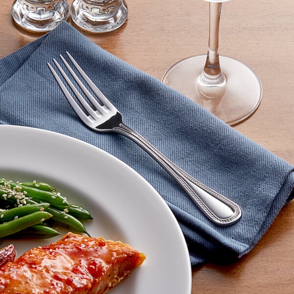 An Acopa stainless steel dinner fork on a plate of food with a napkin and wine glass.