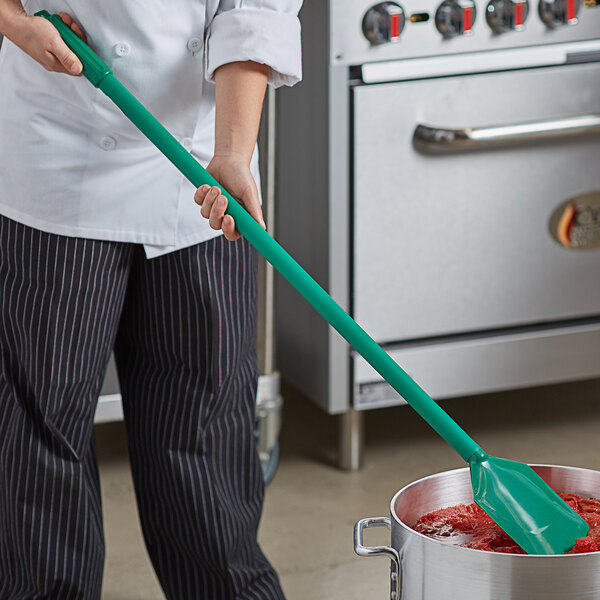 A person holding a Carlisle green paddle with nylon blade and polypropylene handle over a pot of red liquid.