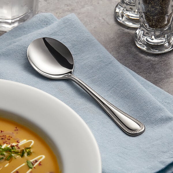 An Acopa Lydia stainless steel bouillon spoon on a napkin next to a bowl of soup.