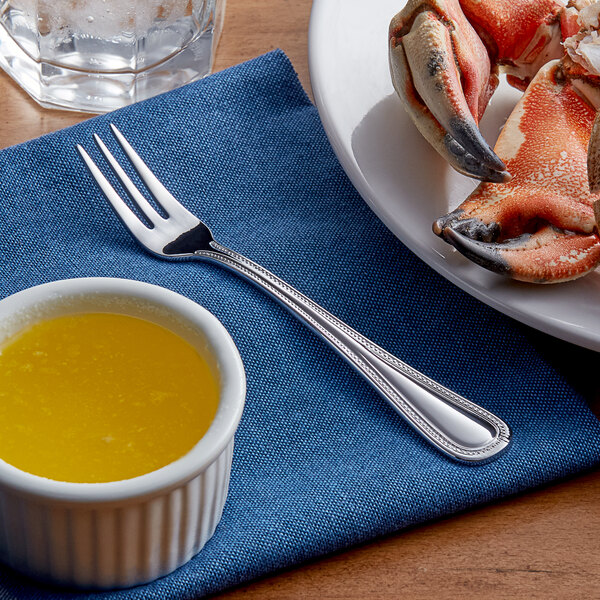 A plate of crab claws with an Acopa Lydia stainless steel cocktail fork and sauce on a blue napkin.