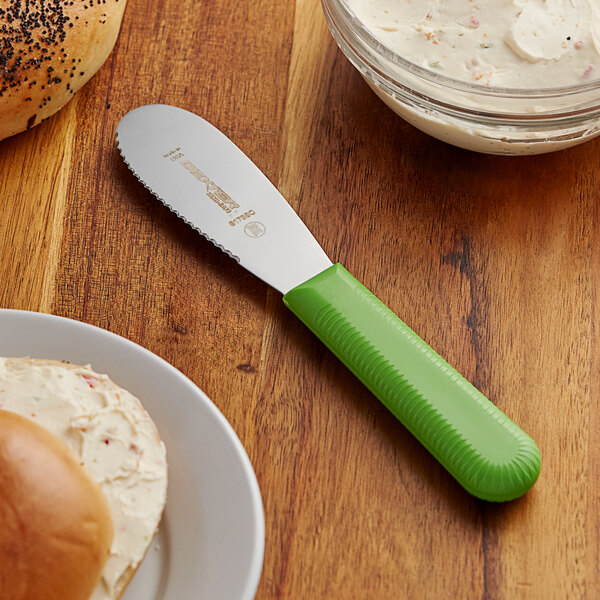 A Dexter-Russell green scalloped sandwich spreader on a table with a bagel.