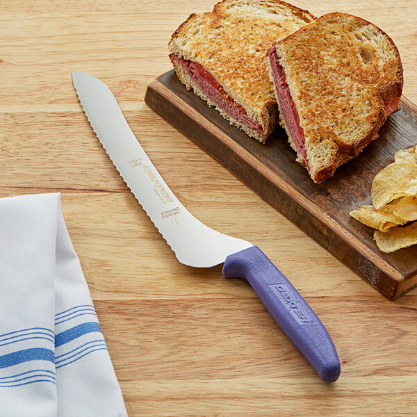 A Dexter-Russell purple scalloped bread knife next to a grilled sandwich on a cutting board.