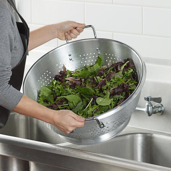 A woman using a Carlisle aluminum colander to rinse greens over a sink.