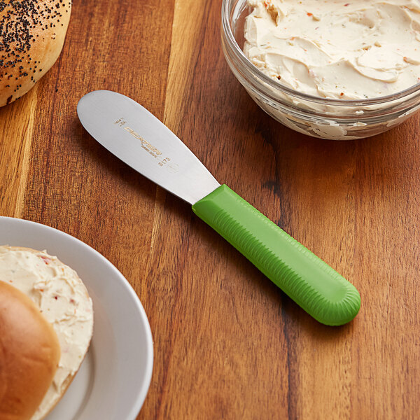 A Dexter-Russell green smooth sandwich spreader on a table with a bowl of cream cheese and a bread.