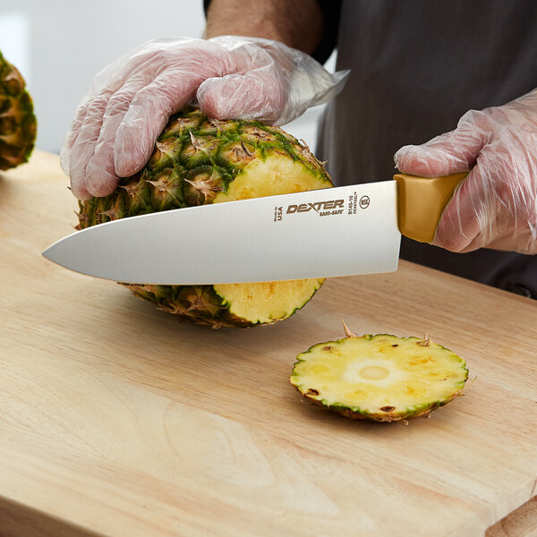 A person using a Dexter-Russell tan chef knife to cut a pineapple on a counter.