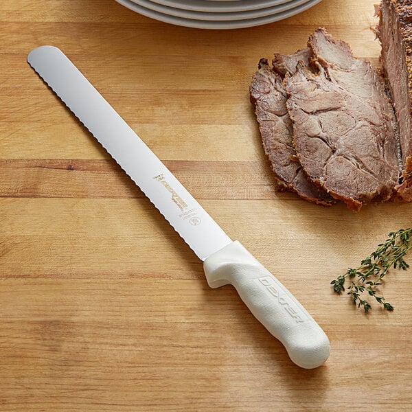 A Dexter-Russell Sani-Safe Narrow Scalloped Slicer with a white handle cutting meat on a table.