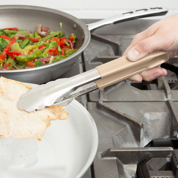 A close-up of a person using Vollrath stainless steel tongs with a tan handle to hold food over a pan.