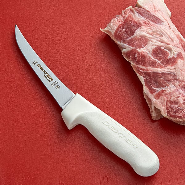 A Dexter-Russell curved boning knife next to a piece of meat on a red surface.