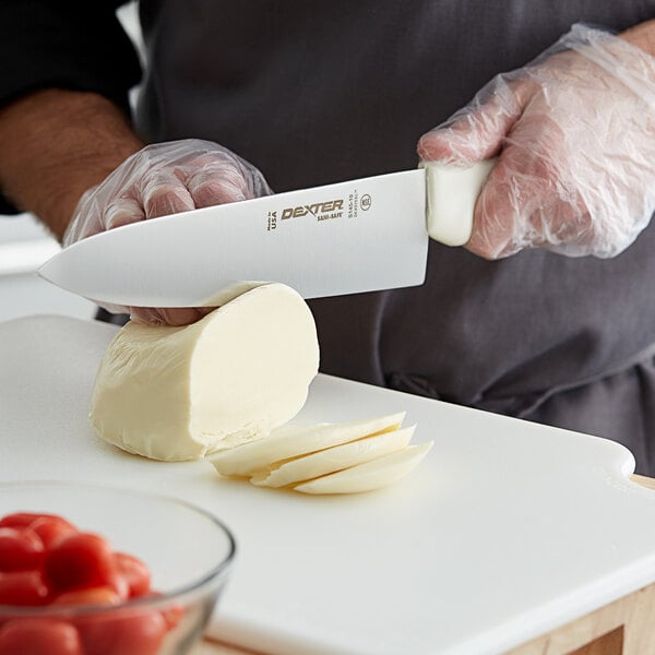 A person using a Dexter-Russell chef knife to cut cheese on a cutting board.