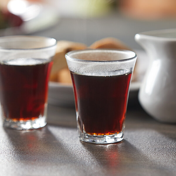 Two Duralex Amalfi shot glasses filled with brown liquid on a table.