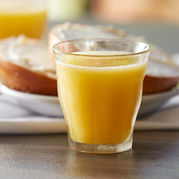 A close-up of a Duralex Picardie stackable glass filled with orange juice on a table next to bread.