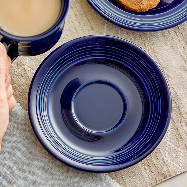 A hand holding a cup of coffee and a blue Acopa Capri stoneware saucer with a plate of food on a table.