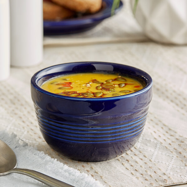 An Acopa Capri deep sea cobalt stoneware bouillon with yellow soup in it on a white cloth with a spoon.