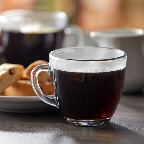A close up of a Duralex glass mug full of coffee on a saucer with cookies.