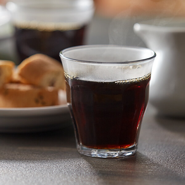 A Duralex Picardie stackable glass filled with dark liquid on a table with steam and a plate of bread.