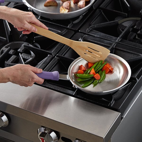 A person cooking vegetables in a Vollrath aluminum fry pan on a stove using a wooden spatula.