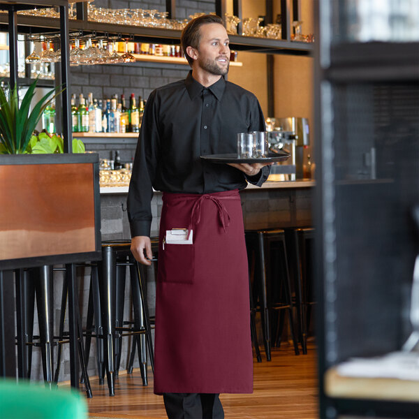 A man in a black shirt and Choice burgundy bistro apron with a pocket holding a tray.