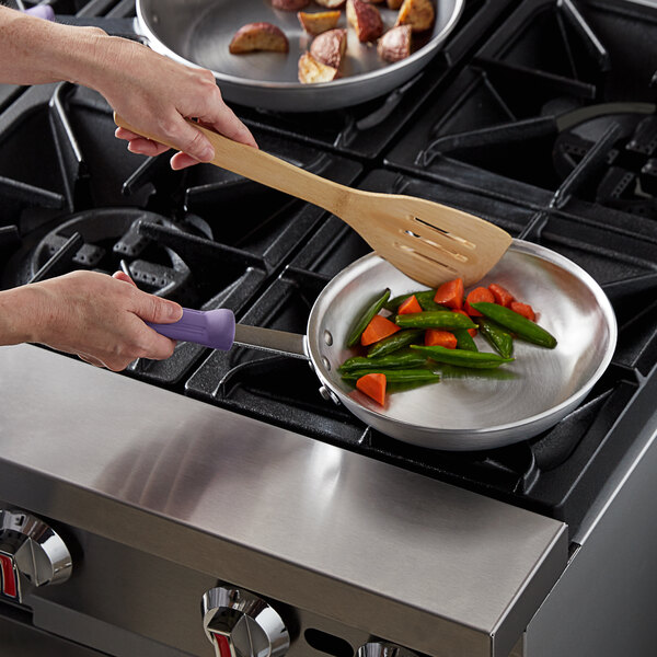 A person cooking vegetables in a Vollrath aluminum fry pan on a stove using a wooden spatula with a purple allergen-free sleeve handle.