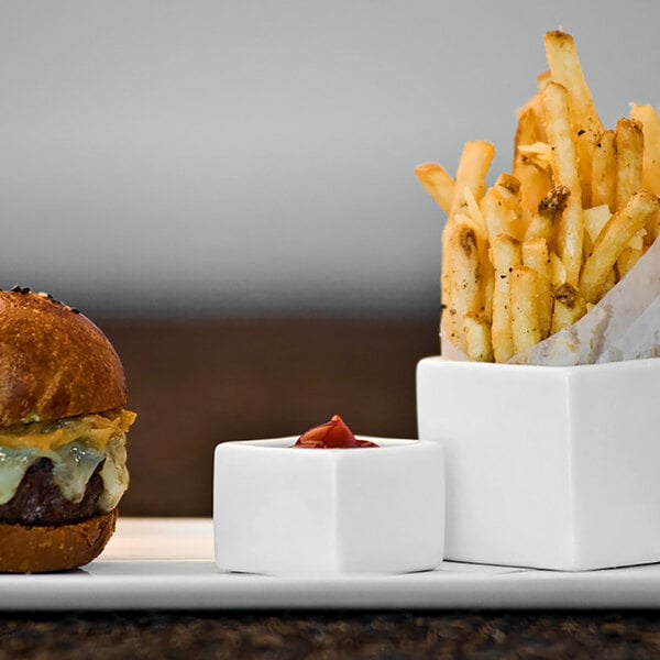 A burger and fries on a plate with a white square porcelain ramekin filled with ketchup.