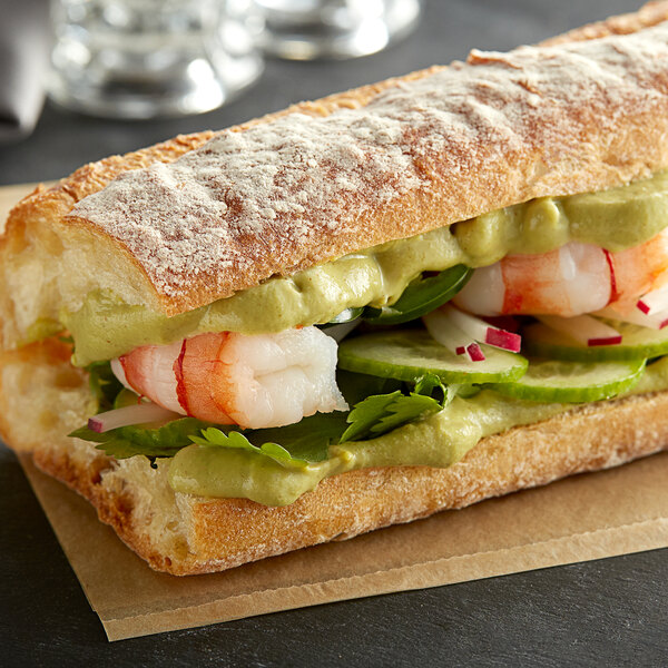A Pilsudski Wasabi Mustard squeeze bottle on a deli counter next to a sandwich with shrimp and avocado.