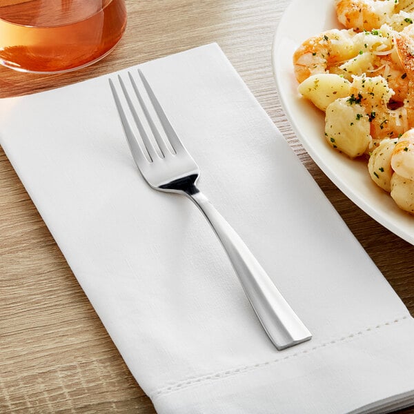 An Acopa stainless steel dinner fork on a white napkin next to a plate of shrimp.