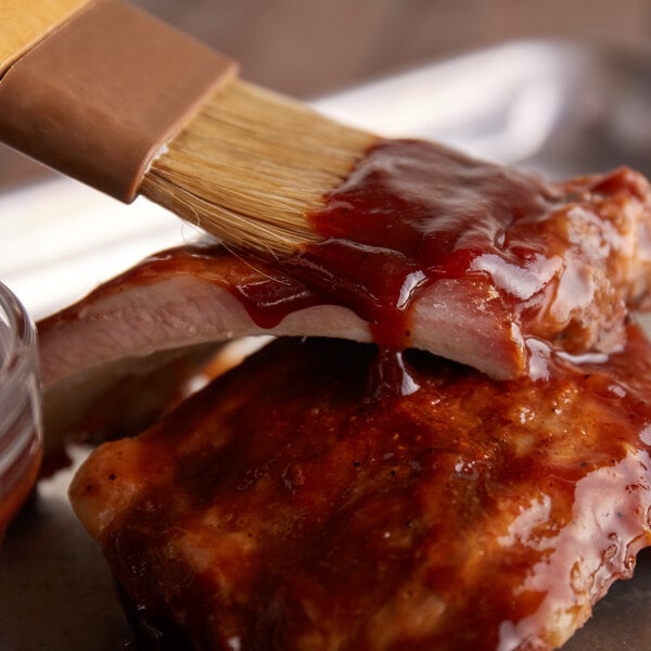A hand using a brush to spread Sweet Baby Ray's BBQ sauce on ribs.