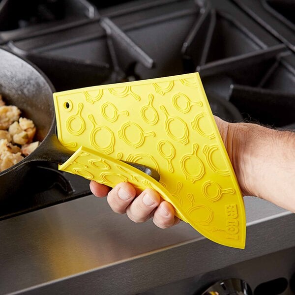 A hand using a yellow Lodge silicone trivet to hold a yellow pan over a pan of food.