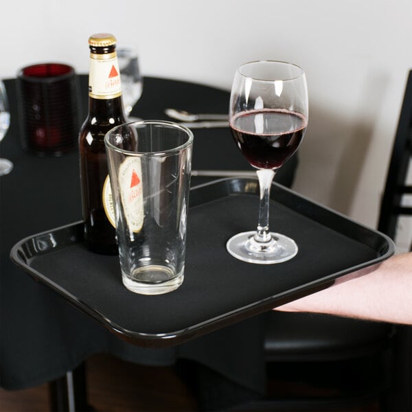 A person holding a Cambro black non-skid serving tray with a glass of wine and a bottle of beer.