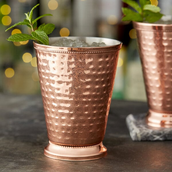 Two Acopa hammered copper mint julep cups on a table with ice and mint leaves.