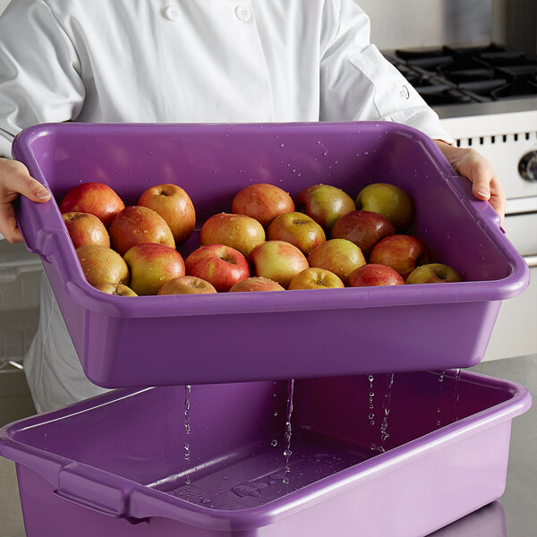 A woman in a white chef's coat holding a Vollrath purple allergen-free drain box full of apples.