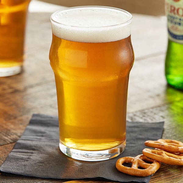 An Arcoroc English pub glass of beer with foam, next to pretzels on a table.