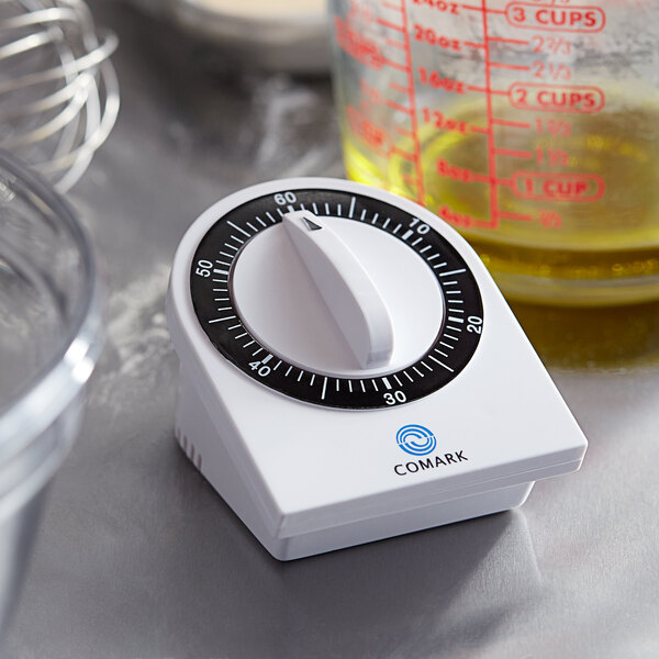 A white Comark kitchen timer with black numbers on a silver surface next to a measuring cup.