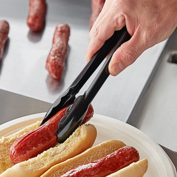 A hand using Visions black tongs to pick up a hot dog.