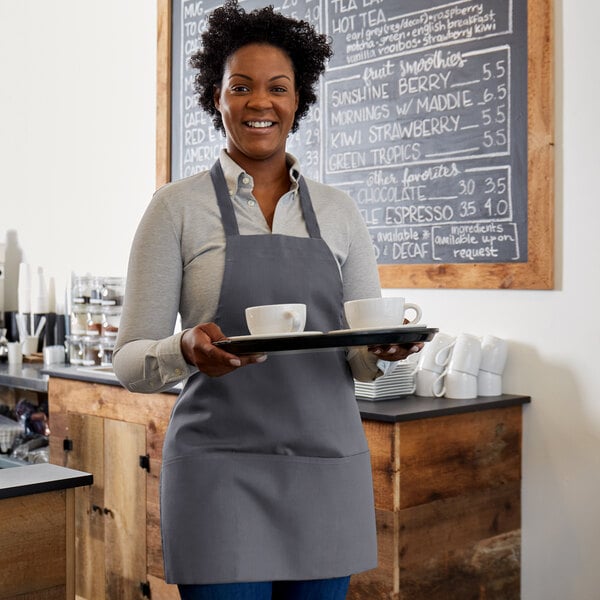 A woman wearing a Choice gray poly-cotton bib apron holding a tray of coffee cups.