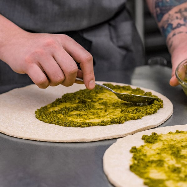 A person using a spoon to spread green pesto on a Rich's Fresh N Ready pizza crust.