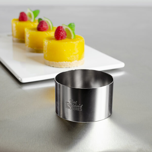 A stainless steel round food ring mold with a round object on it next to a plate of desserts.